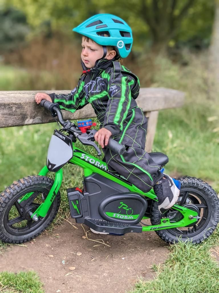  small boy wearing a green and black wulfsport suit, sitting on a green 12" storm electric balance bike.